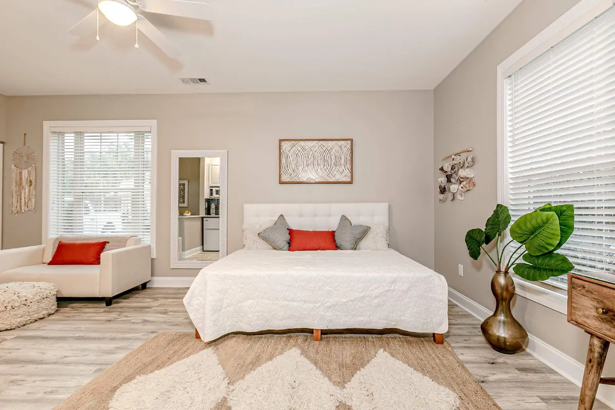 A tranquil bedroom with white bedding, decorative pillows, a large mirror, indoor plant, and soft natural light filtering through the windows. The ambiance suggests a peaceful retreat ideal for unwinding together.