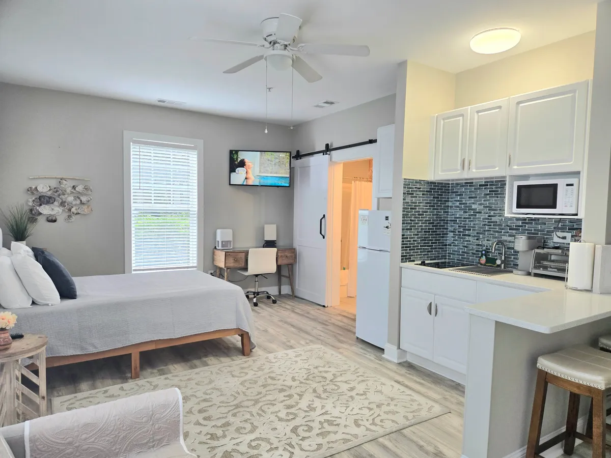 This image presents a cozy and functional studio layout with a plush bed, a streamlined kitchenette, a compact desk area, and a television. Natural light streams in, illuminating the practical yet welcoming space.