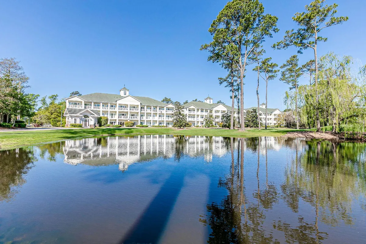An inviting exterior image of the resort building, reflected in a tranquil pond and surrounded by tall trees. The peaceful landscape is perfect for leisurely strolls, outdoor relaxation, and appreciating the resort’s serene setting.