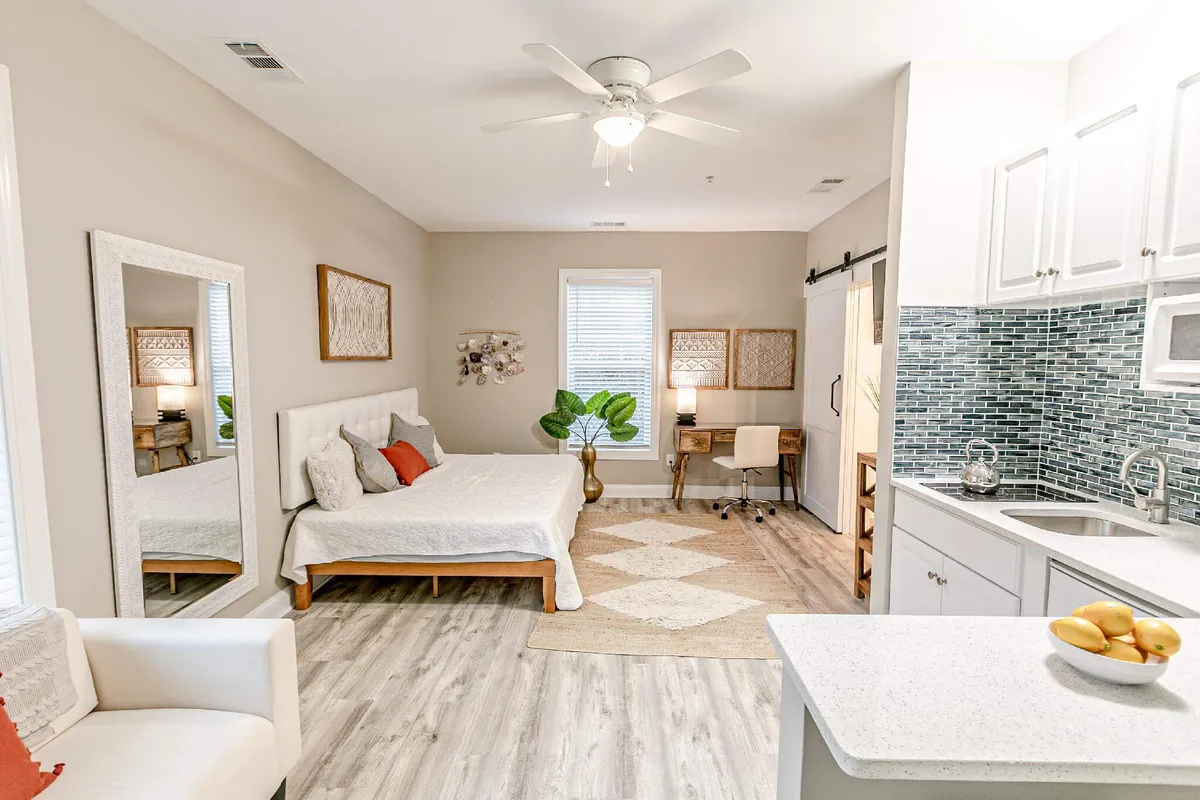 A stylish studio space featuring a comfortable bed, a sleek kitchenette with modern appliances, a mirror, a small desk, and a leafy plant. The cohesive design makes the space feel both airy and inviting.