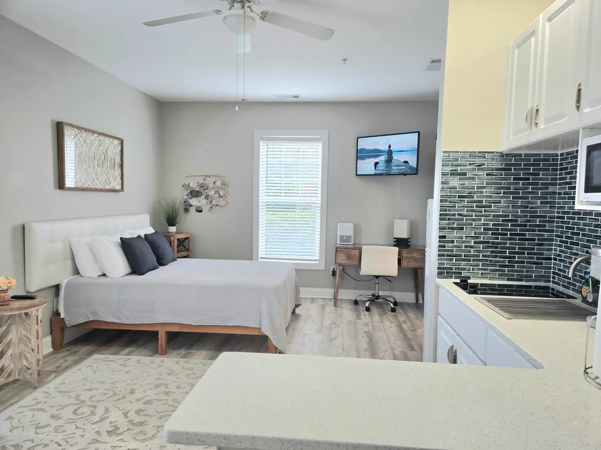 This image showcases a bright, inviting studio with a bed adorned with decorative pillows, a window letting in ample natural light, a workspace with a chair, a television displaying a beach scene, and tasteful wall art. The overall ambiance is serene and comfortable, suggesting both relaxation and productivity.