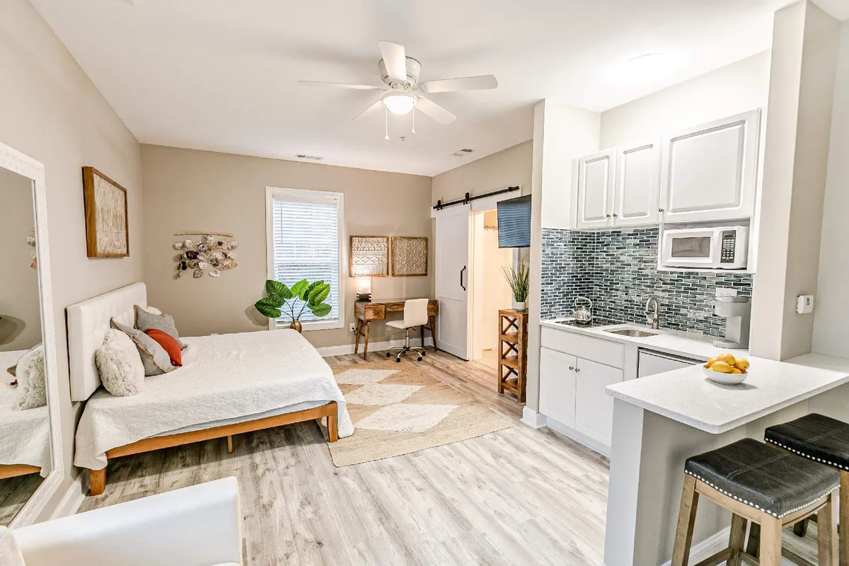 The scene highlights a bed with crisp bedding, a compact dining or desk area, a kitchenette, a decorative mirror, and greenery. Soft morning light enhances the sense of comfort and functionality.