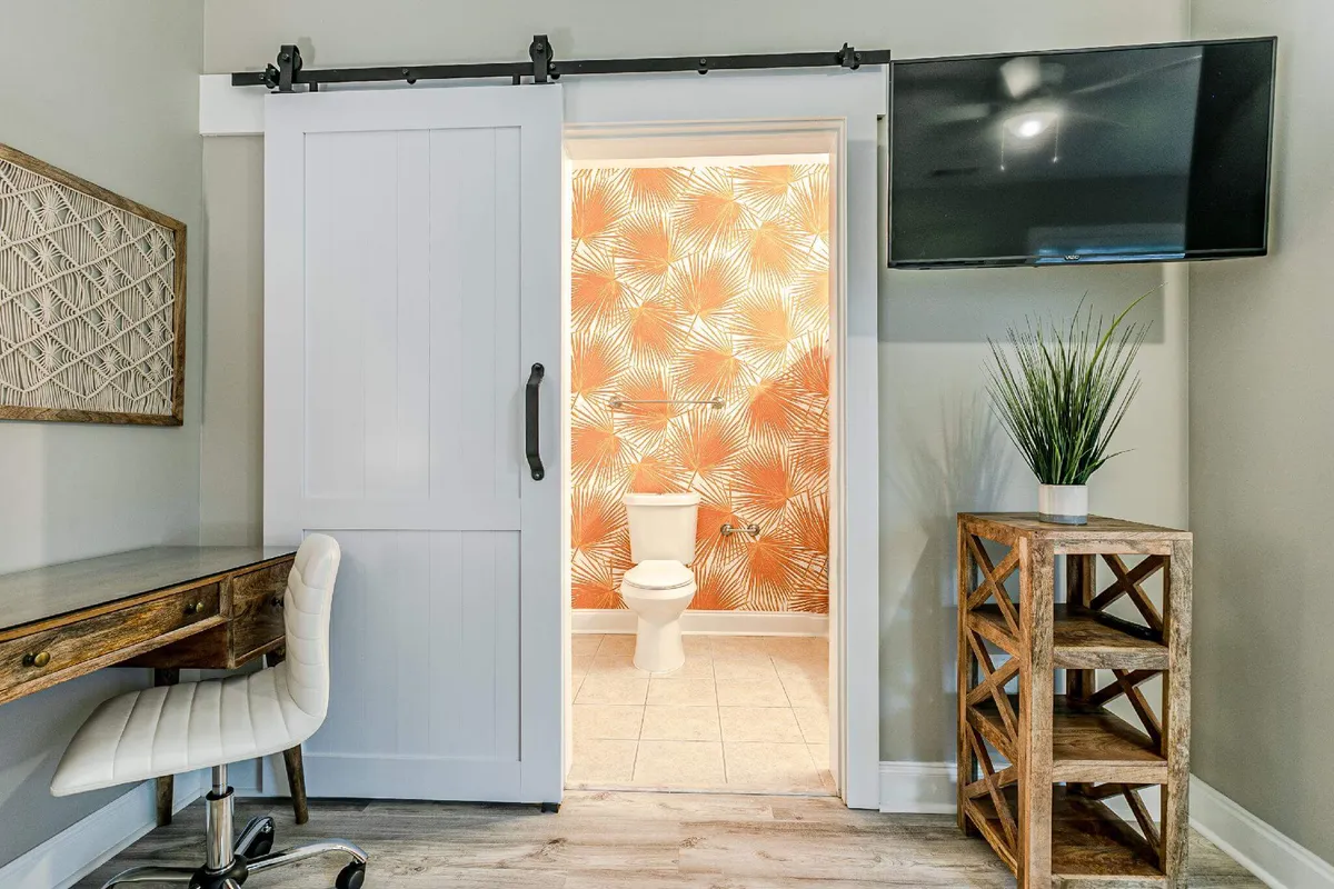 A modern, cozy nook designed for productivity, showcasing a wooden desk with a chair beside a sliding barn door. The area is decorated with wall art, a TV, and a touch of greenery, creating an inviting spot ideal for remote work or focused tasks. The bathroom entrance is subtly visible, suggesting the convenience of having everything close at hand.