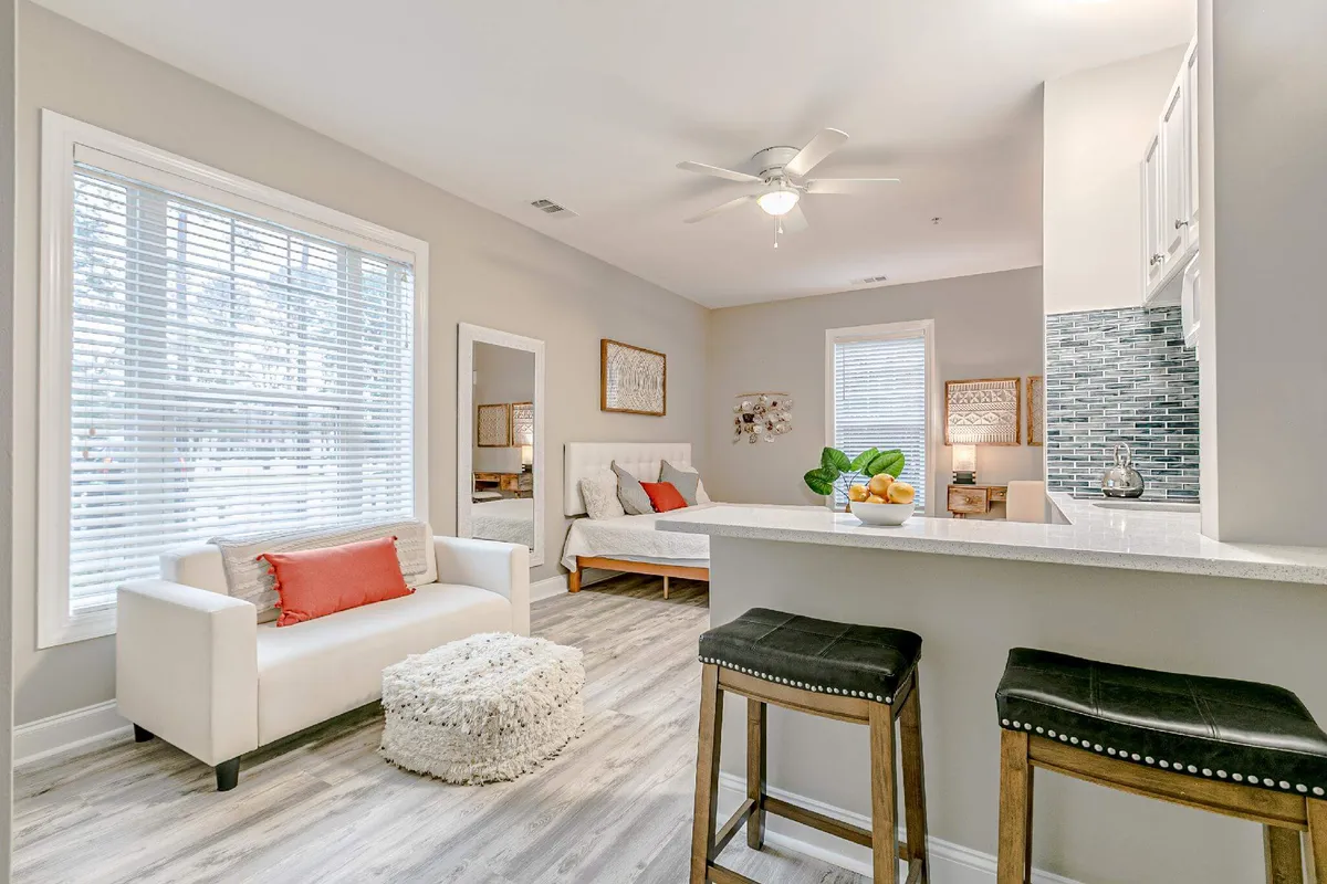 The image showcases a stylish, sunlit studio interior featuring a cozy sofa with a vibrant red accent pillow, a sleek kitchen bar topped with a fresh fruit bowl, and tasteful wall art. Morning light pours through the windows, illuminating the welcoming space designed for relaxation and intimate dining.