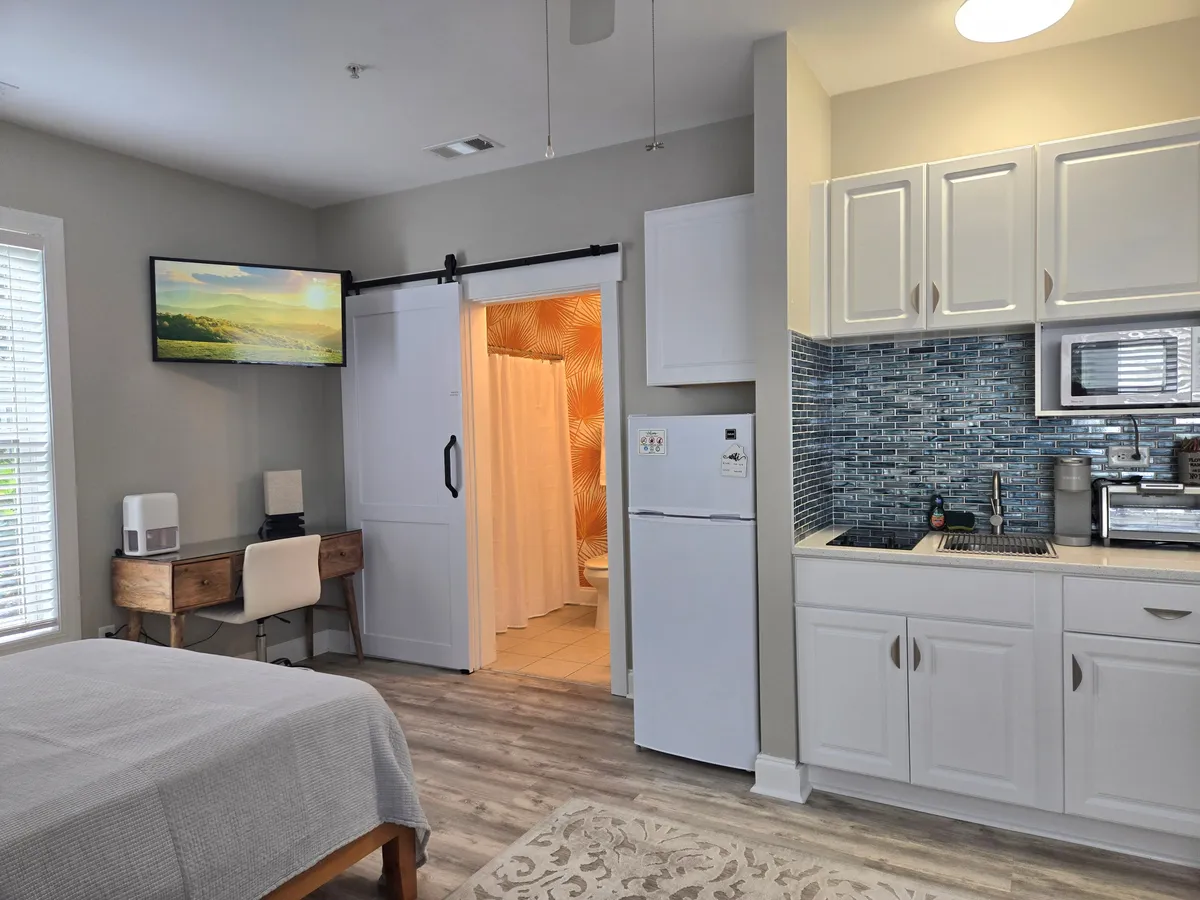 This image offers a wide view of the studio’s interior, including a glimpse of the shower area through the visible shower curtain. The photo captures parts of the bed, desk, TV, kitchen, and importantly, the bathroom zone, conveying the functional and inviting ambiance of the private bath space within the larger studio context.