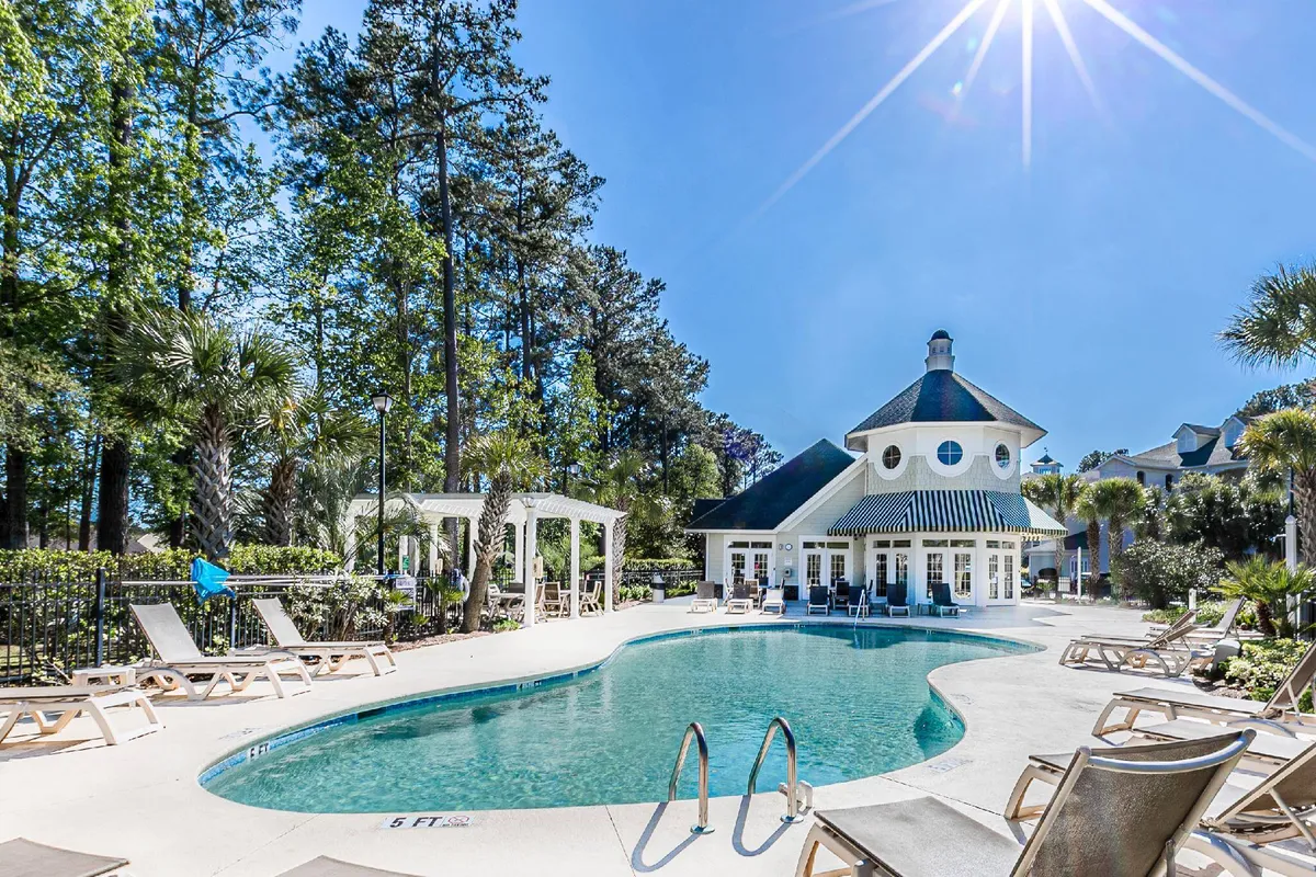 This exterior image captures a sparkling swimming pool surrounded by lounge chairs and palm trees, with a view of the clubhouse in the background. The scene features people enjoying the water and relaxing poolside, evoking a vibrant resort atmosphere.