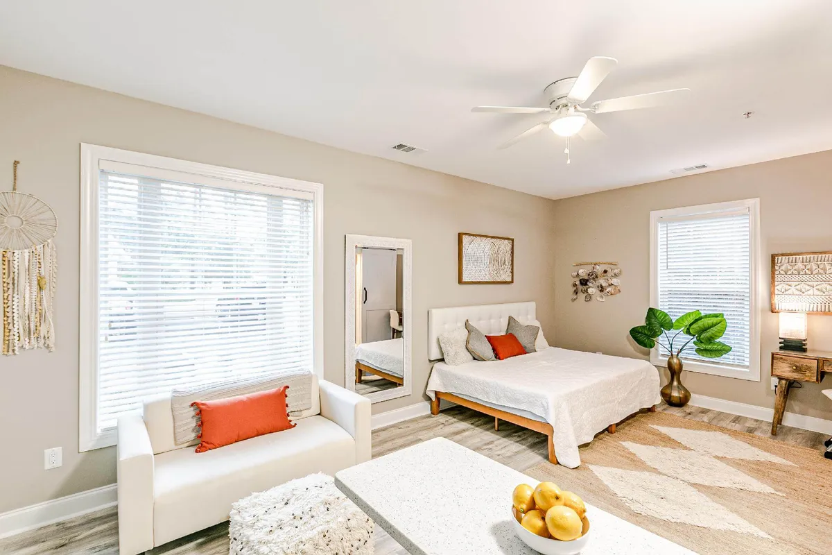 A serene corner of the studio featuring a white bed with decorative pillows, a cozy sofa with a bright orange accent pillow, decorative wall art, and a lush green indoor plant. The scene is bathed in gentle natural light, creating a peaceful, welcoming environment.